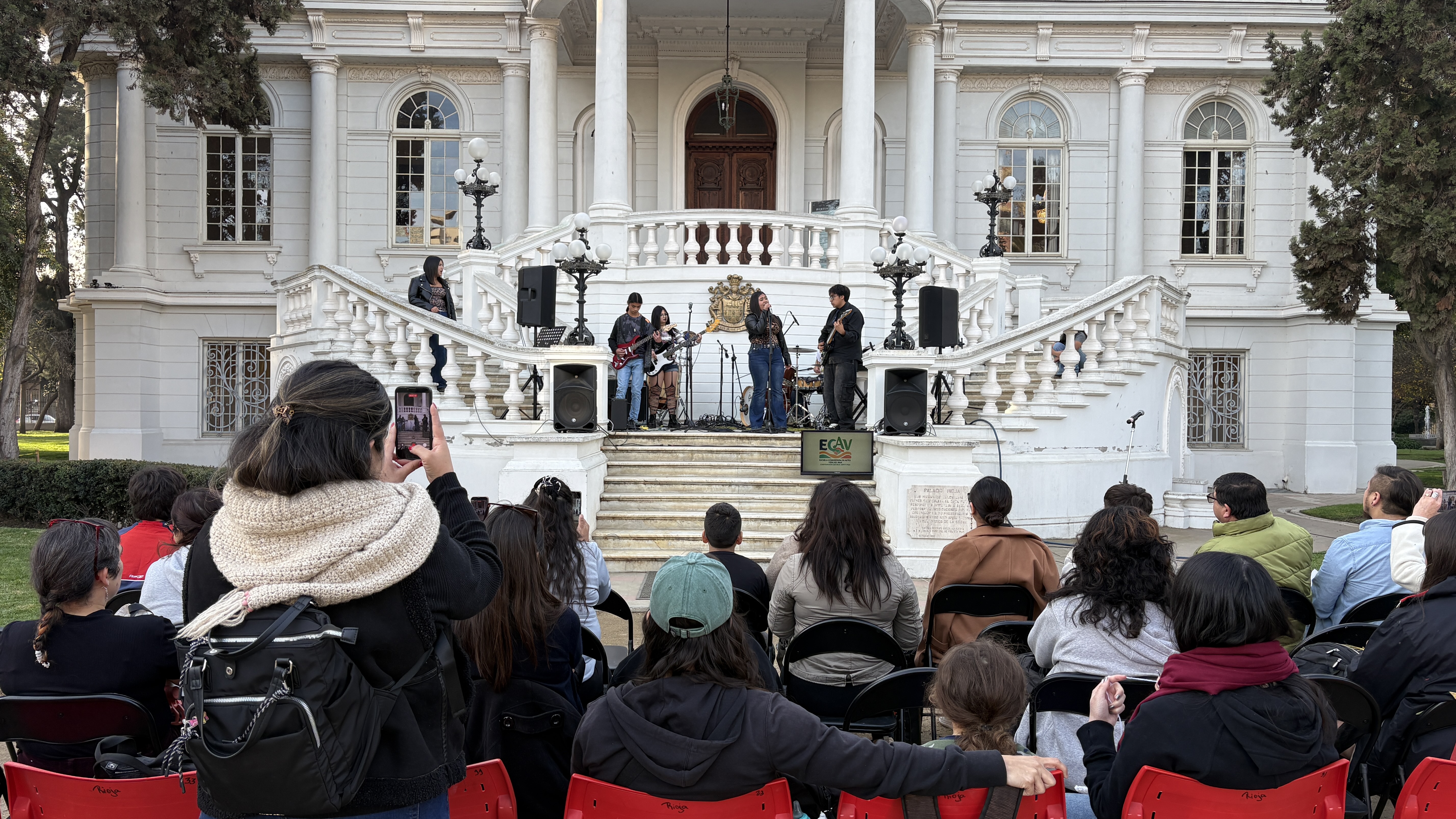 Presentación de estudiantes de ECAV frente a un edificio patrimonial y público asistente.